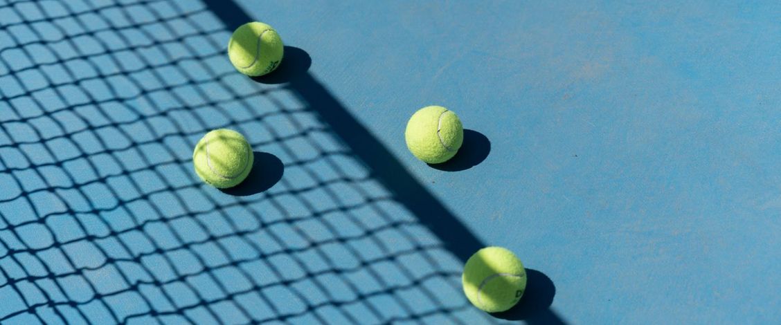 Four yellow tennis balls resting on a bright blue hard court in sunshine, with the shadow of the net falling diagonally across the surface