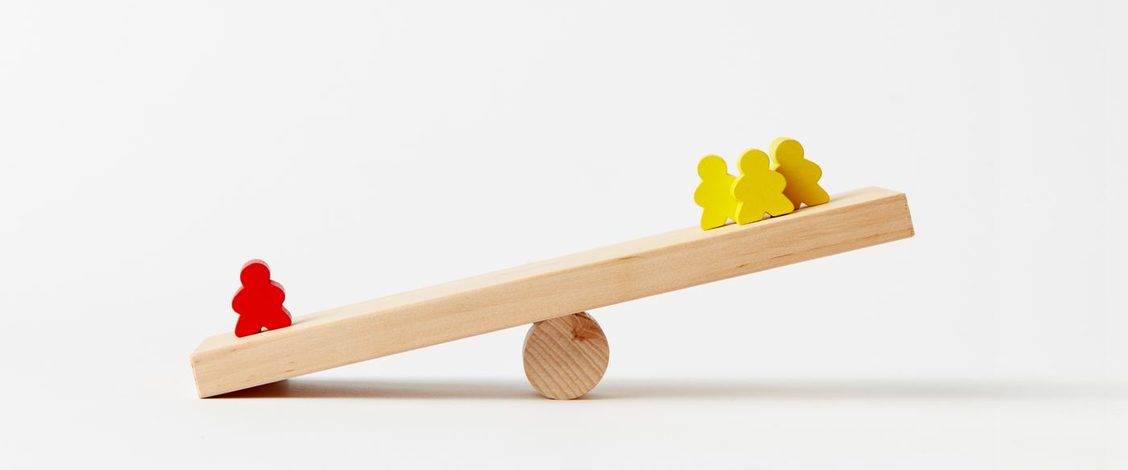 A plank of pale wood balanced on a round wooden block, tipped down at the left end where one red peg figure stands alone, while three yellow peg figures stand together at the raised right end, against a plain white background