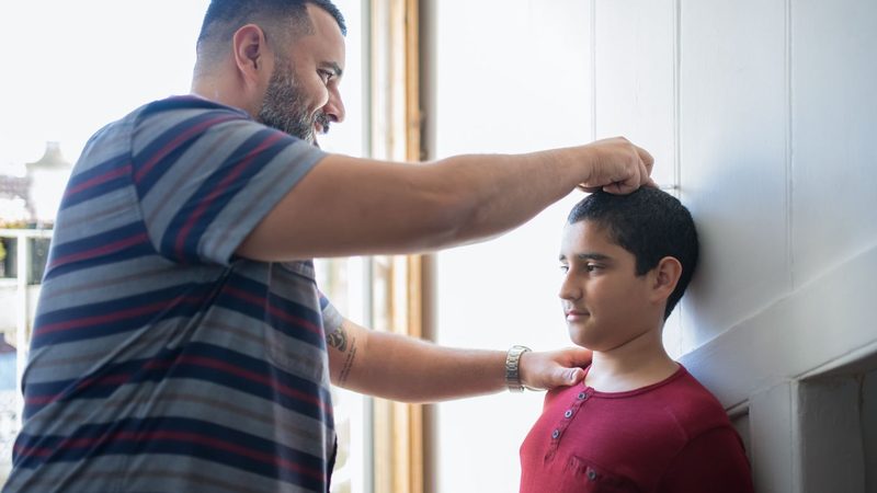 A father in a striped T-shirt marks his son&rsquo;s height against a white wall in a sunlit room while the boy stands straight with his back to the wall