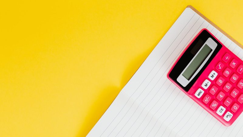 A pink calculator lying on an open lined notebook against a bright yellow background