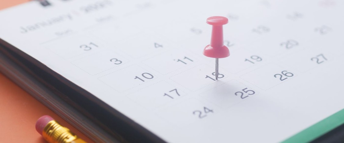 A pink push pin stuck into one date on a calendar page, with a yellow pencil and two paper clips on an orange background.