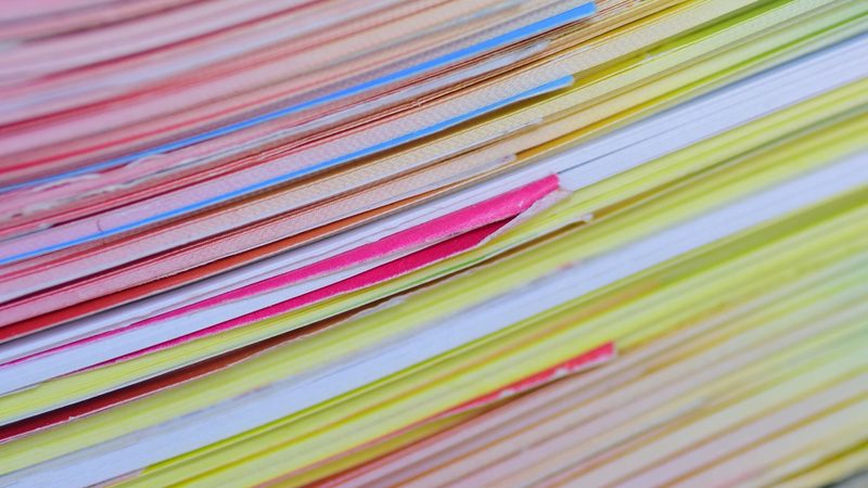 The edges of a tall stack of coloured papers and folders in pink, yellow, blue and white, seen close up and side on