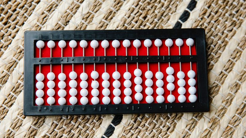 A black abacus with white beads on a red board, lying on a woven straw mat