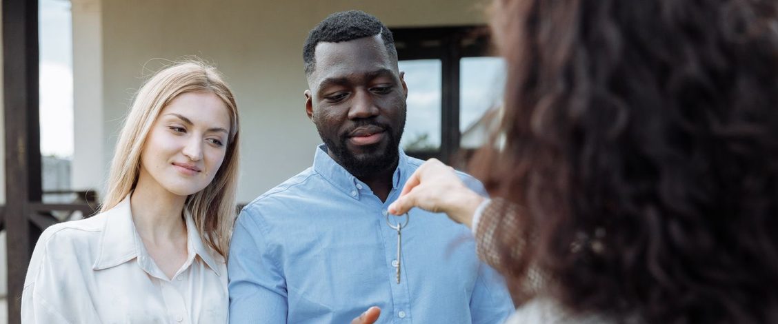 A woman with long dark curly hair, seen from behind, holds out a silver house key on a ring to a smiling couple standing arm in arm outside a house in daylight, the man in a pale blue shirt reaching out his open hand to take it and the woman beside him in a cream blouse watching